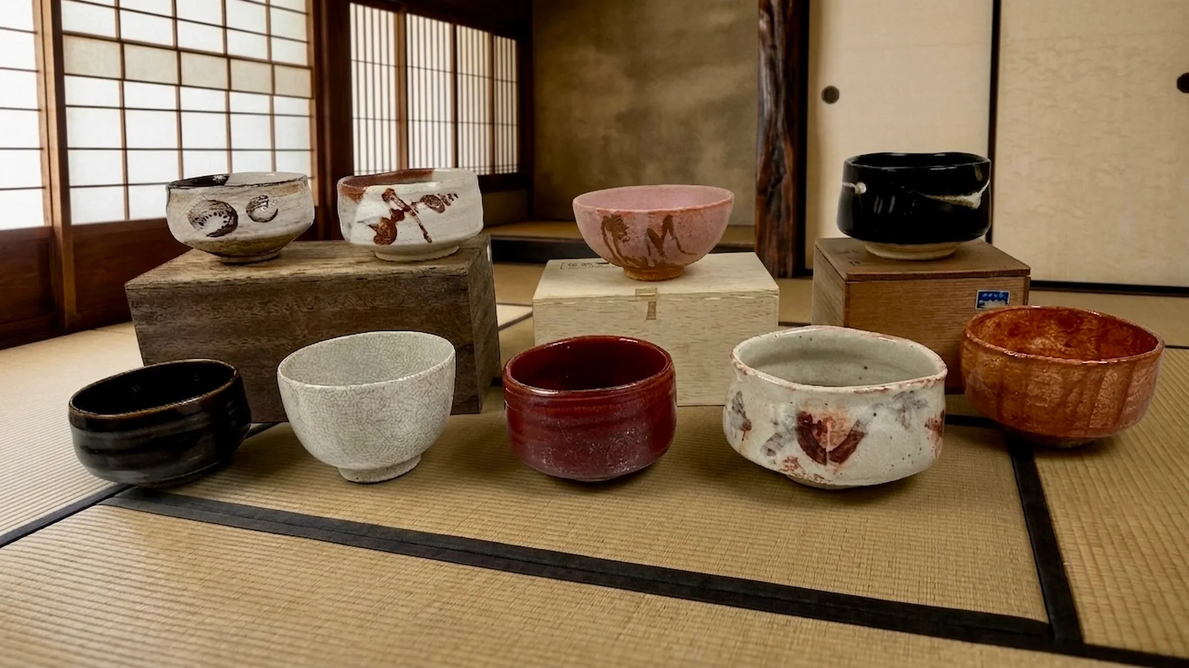 A curated collection of eight antique Japanese tea bowls (chawan) and traditional paulownia wooden storage boxes (kiribako) displayed on a tatami mat floor inside a rustic, dimly lit Japanese tea room (chashitsu) with shoji paper screens.